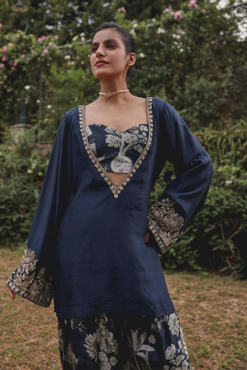 Shajar three-piece festive set styled in a rose garden — navy cotton modal kurta with gold embroidery over tree-of-life printed bralette and farsi salwar